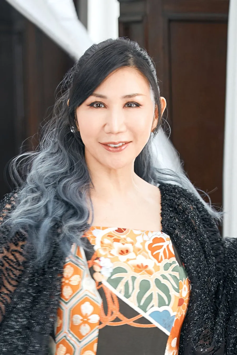 女性のポートレート。黒髪に青みがかったグレーの髪が混ざり、黒いトップスとオレンジと緑の柄の着物風の服を着ている。彼女は微笑んでおり、顔は化粧をしている。