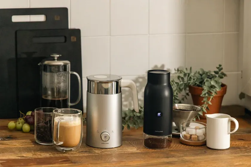 コーヒーメーカー、コーヒー豆、砂糖、ミルクピッチャー、コーヒーカップ、ブドウ、観葉植物などが木製のテーブルに置かれた写真。