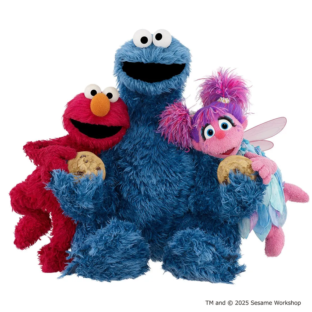 セサミストリートのキャラクター、エルモ、クッキーモンスター、アビー・カダビーが寄り添っている画像です。エルモとアビーはクッキーを手に持っています。