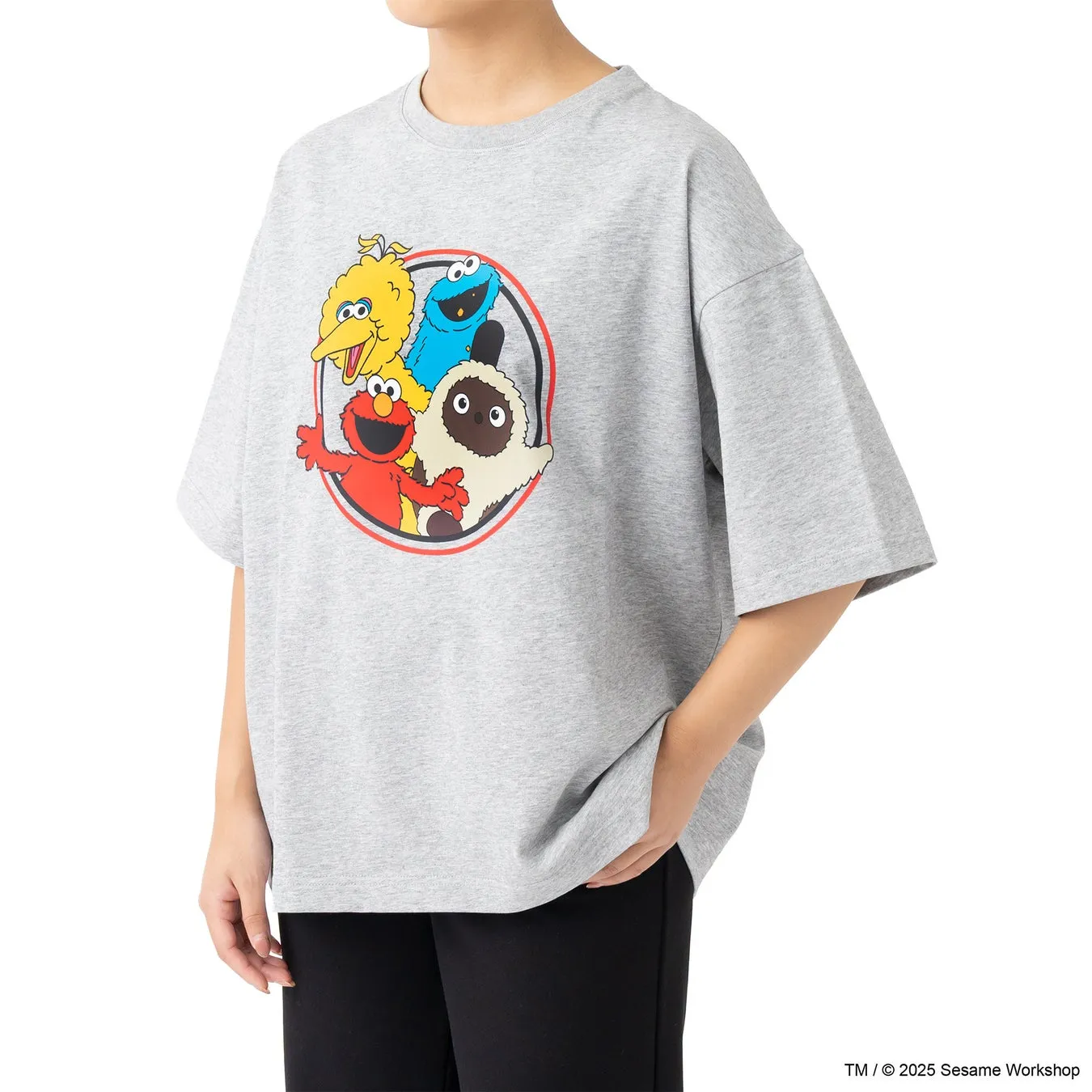 グレーのTシャツを着た女性が立っている画像。Tシャツにはセサミストリートのキャラクターが描かれており、丸いデザインの中にエルモ、クッキーモンスター、ビッグバード、そしてモッピーが描かれている。